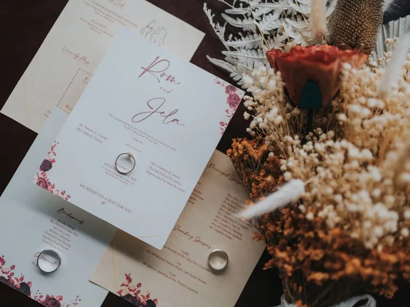 A wedding invitation suite laid flat with two gold rings and a dried-flower bouquet.